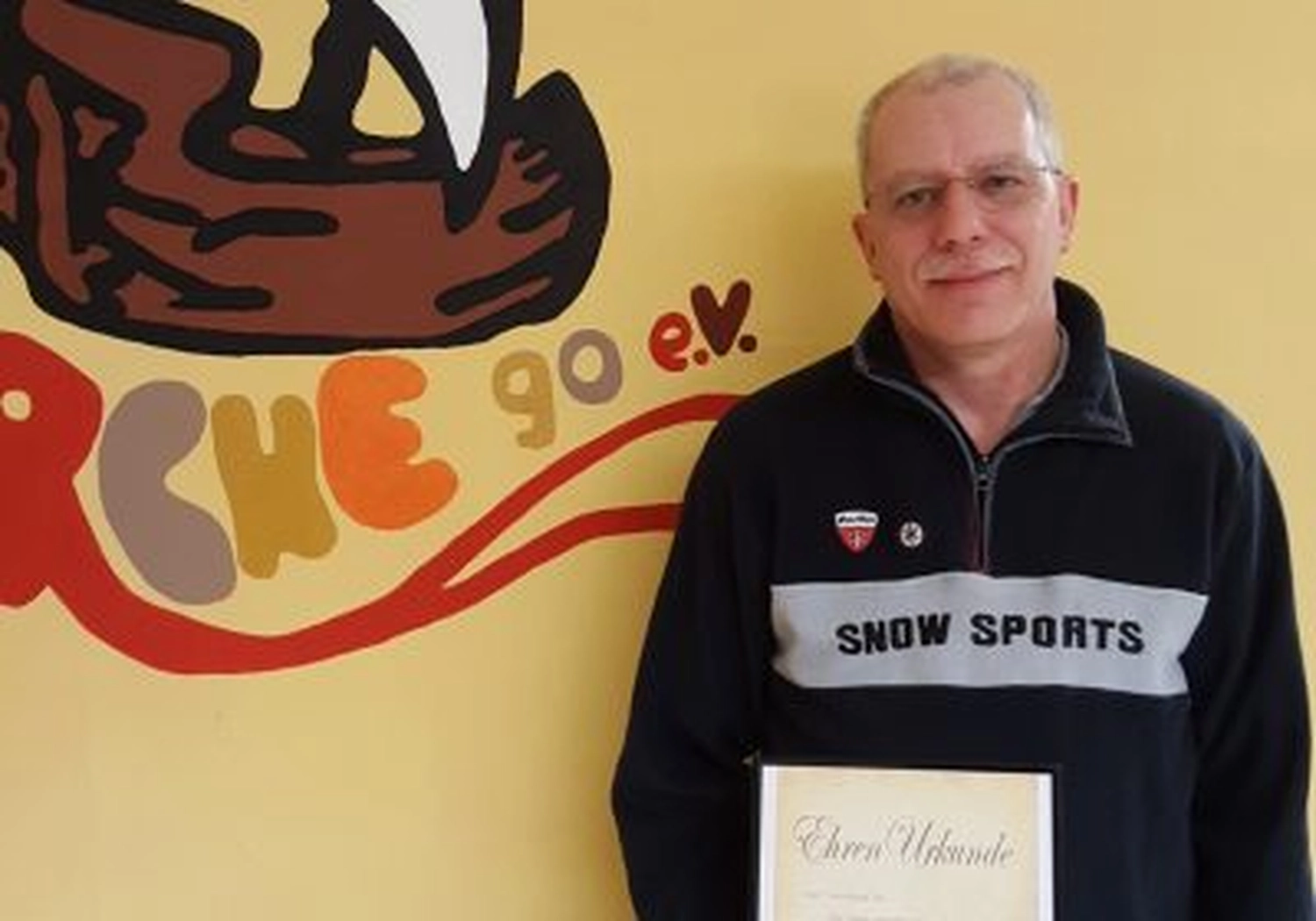 Tierschutzorganisation Arche 90 e.V. - Ein Mann in einem Pullover mit der Aufschrift „Snow Sports“ steht neben einem gelben Logo mit der Aufschrift „CHE 90 e.V.“ und hält eine gerahmte Urkunde.