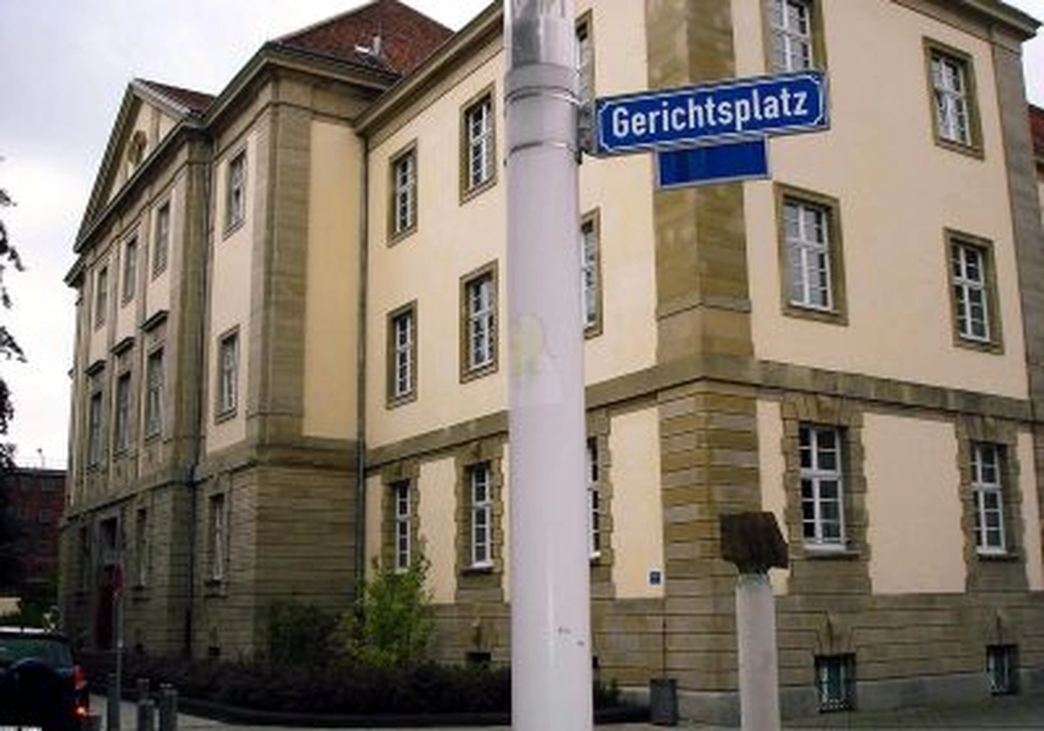 Tierschutzorganisation Arche 90 e.V. - Ein klassisches europäisches Steingebäude unter blauem Himmel, teilweise verdeckt von einem Straßenlaternenmast mit einem blauen Schild „Gerichtsplatz“.