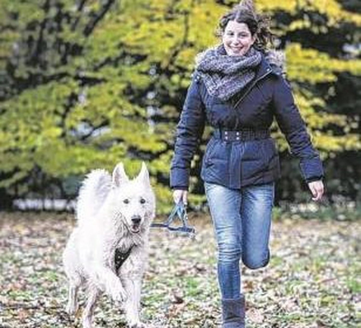 Tierschutzorganisation Arche 90 e.V. - Eine Frau in Winterjacke und Schal lächelt, während sie mit einem flauschigen weißen Hund an der Leine durch einen Park mit Herbstlaub spaziert.