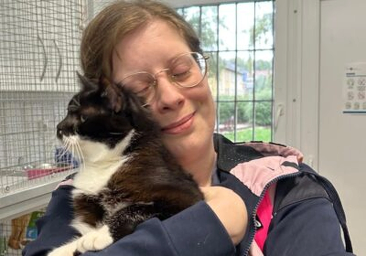 Tierschutzorganisation Arche 90 e.V. - Eine Frau mit Brille kuschelt mit einer flauschigen, schwarz-weißen Tuxedo-Katze in einem Raum mit Käfigen.