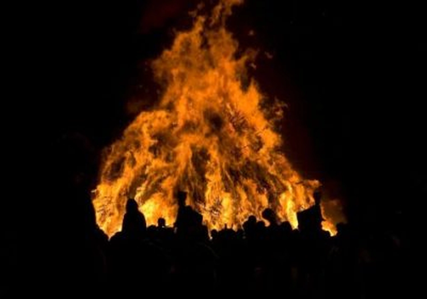 Nachtaufnahme eines großen, lodernden Lagerfeuers mit intensiv orangefarbenen und gelben Flammen vor pechschwarzem Hintergrund.