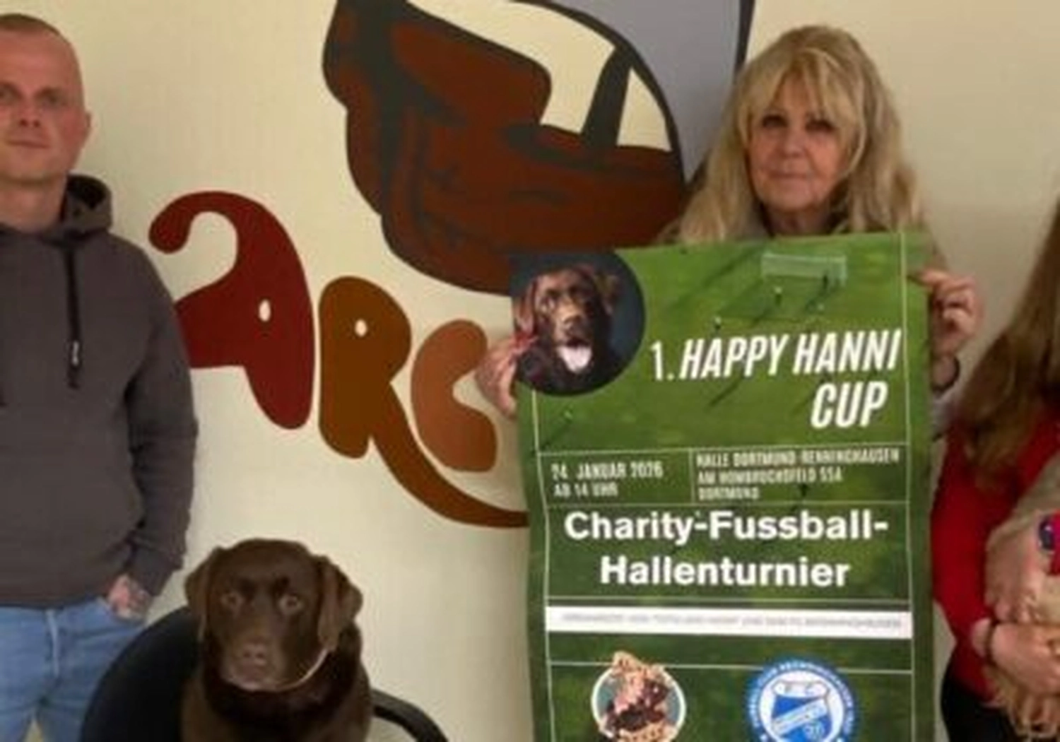 Tierschutzorganisation Arche 90 e.V. - Eine Gruppe von Personen steht in einem Innenraum neben einem schokoladenbraunen Labrador Retriever und hält ein großes grünes Werbeplakat hoch.